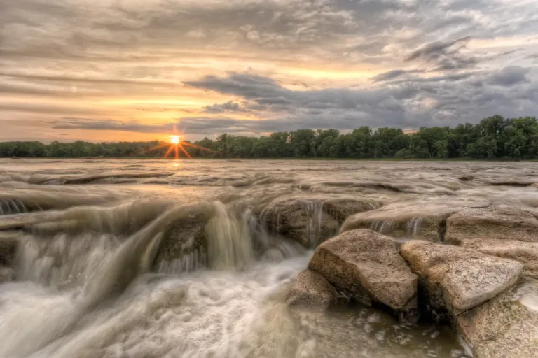 Rushing water over large river rocks with a vibrant orange sunset in the sky at Otsego Park.